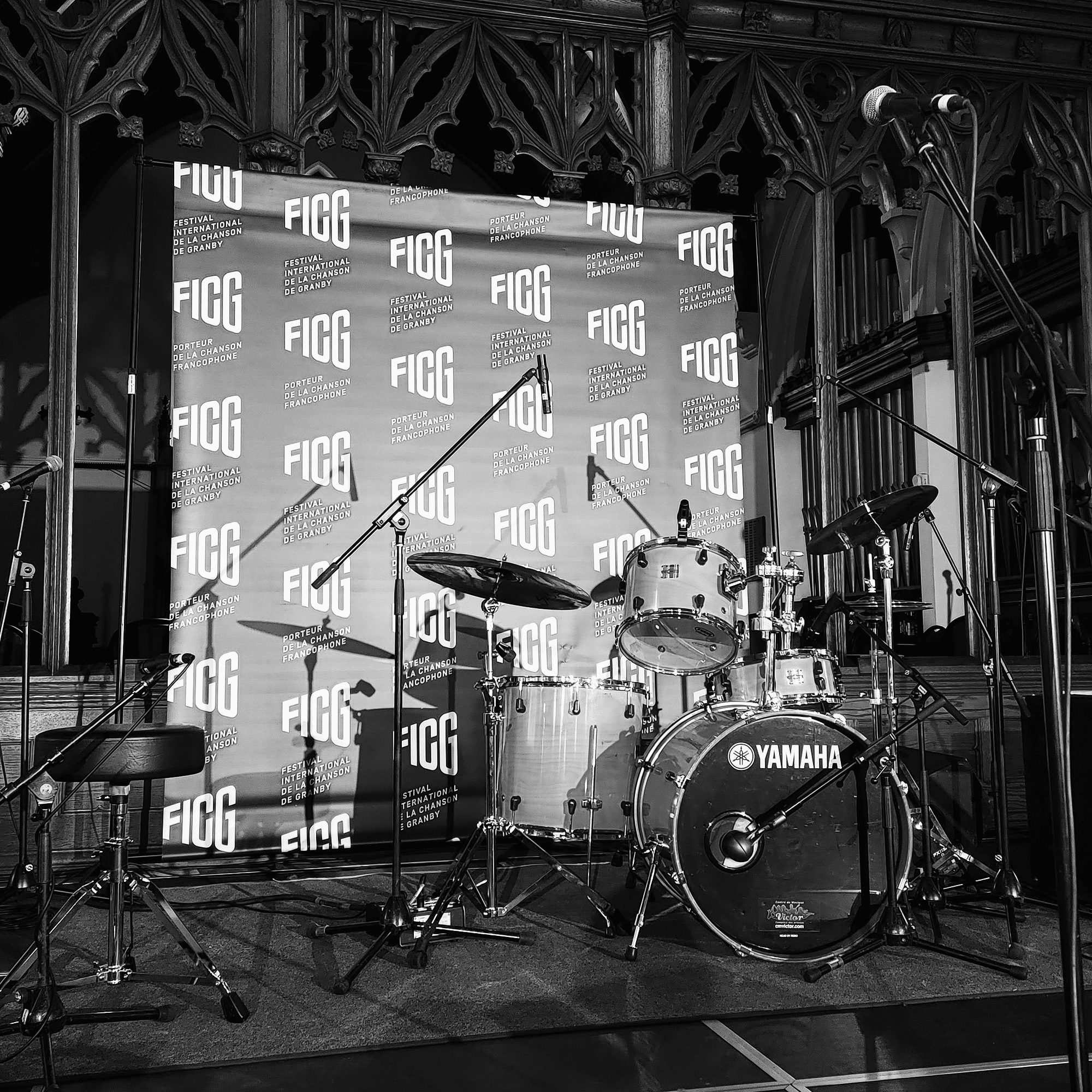 Une photo en noir et blanc d'une batterie sur scène, comprenant une grosse caisse Yamaha et d'autres instruments de percussion. Plusieurs microphones se trouvent autour. La toile de fond indique « FIGG » à plusieurs reprises. L’arrière-plan présente des détails en bois ornés.