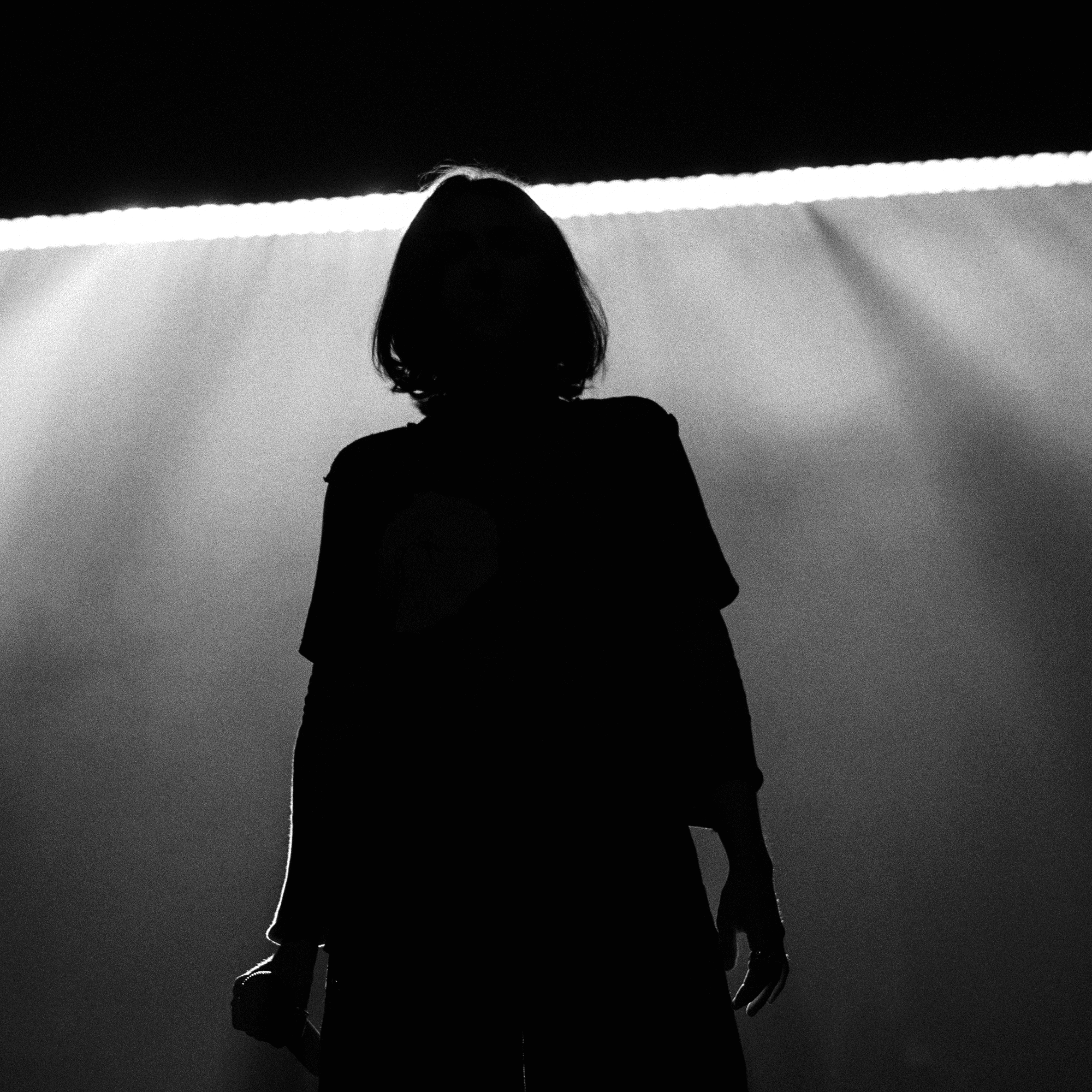 Une photo en noir et blanc montre une silhouette se tenant devant une source de lumière vive, créant des ombres spectaculaires. La lumière est positionnée derrière l’individu, obscurcissant les détails et créant un fort effet de rétroéclairage.